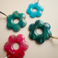 Collection of daisies, one is vibrant pink, a dark teal, and a brighter teal.  These all have specks of golden glitter.  There is a smaller ice blue daisy as well.