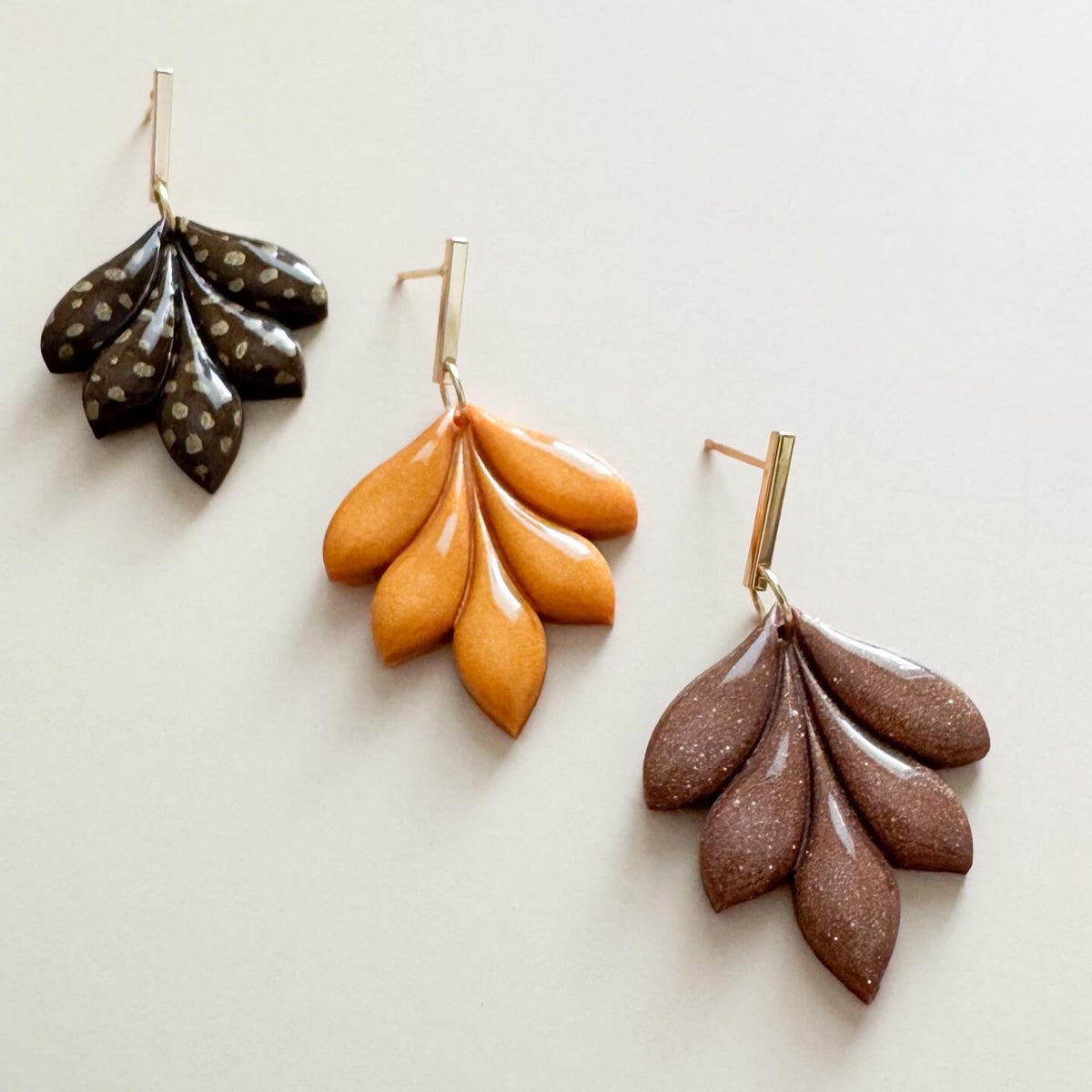 Three pairs of leaf-shaped earrings in brown, gold, and fawn on a light background