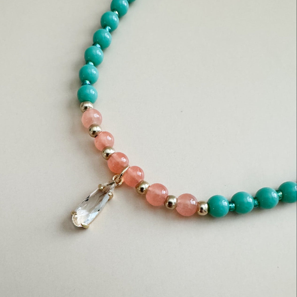 Beaded necklace with mint green and translucent orange beads with an elongated teardrop gem on a light gray background