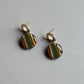 A pair of gold-tone stud earrings with circular drop charms featuring vertical stripes of olive green, gold glitter, and brown on a light gray background.