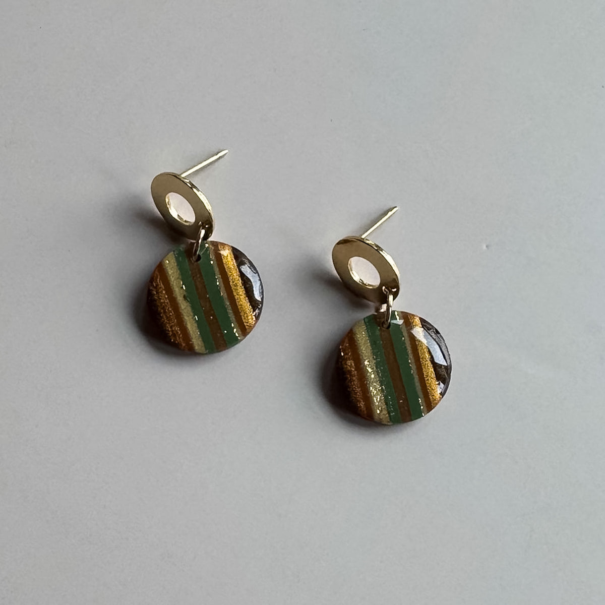 A pair of gold-tone stud earrings with circular drop charms featuring vertical stripes of olive green, gold glitter, and brown on a light gray background.